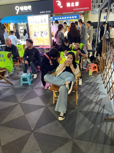 The image shows a street scene in Hang Zhou Shi, China, at the coordinates 30.264 latitude, 120.155 longitude. There are several people in the scene, including a young woman wearing blue jeans and a black shirt sitting on a wooden chair, a young man seated beside her wearing a black shirt, and several other people standing in the background, some of whom are wearing yellow shirts. There are also various objects in the scene, including a street sign, a metal barrier, a folding chair, and a variety of other items. The environment is a city street with a tiled floor and a street sign that reads "No. Nine Shoes Club." The people are interacting with each other. The image was captured from a slightly elevated angle, providing a clear view of the scene.