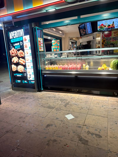 The image shows a food stand on a sidewalk in Sant Antoni De Portmany, Spain. The stand has a large illuminated menu board on the left displaying "APERITIVI PER TUTTI I GUSTI" and images of various fried food items and drinks. To the right, a glass display case exhibits an assortment of colorful fruit cups and a large watermelon. Above the display case, digital screens show menu items including "APPETIZERS", "PIZZA", and "NATURAL JUICES". The sidewalk in front of the stand is paved with square tiles and shows signs of wear and tear, with some stains and debris scattered across it. The overall scene suggests a commercial area, likely a street or promenade, catering to tourists or locals seeking quick food and drinks.