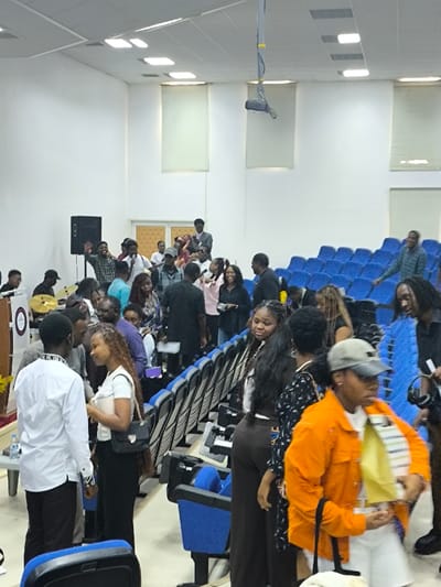 The image depicts an indoor gathering of approximately 30-40 individuals, primarily young adults, within an auditorium or lecture hall in Kızılbaş, Northern Cyprus / Cyprus. The room features white walls, a white ceiling with recessed rectangular lights, and light-colored roller blinds covering windows or panels on the upper walls. A projector is suspended from the ceiling in the upper-middle frame.
In the foreground and mid-ground, attendees are standing and conversing in aisles between rows of blue, cushioned auditorium seats, some of which are occupied. On the left side of the image, a speaker stand with a visible emblem is positioned next to a partial drum set showing cymbals, and a large black speaker is visible against the wall. A small table with bottled water is present near the podium.
Individuals are observed in various postures: some are standing and interacting, such as a woman with long braids conversing with a man in a white long-sleeved shirt on the left mid-ground. Another person in an orange jacket and a light-colored baseball cap is partially visible in the foreground right, holding a small package or item. In the background, center-left, a group of people is gathered near the wall, with some individuals raising their hands, and one person appears to be holding a microphone. A light-colored door is visible in the background, center. A man in a blue plaid shirt is standing in an aisle on the right, smiling. The general atmosphere suggests an event, possibly an assembly, lecture, or social gathering.