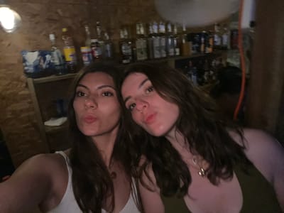 Two women are positioned closely together, with their faces dominating the frame. Behind them is a bar with numerous bottles of alcohol and other items arranged on shelves. The bar appears to be located in a dimly lit area with wooden walls and floors. The location is Puerto Escondido, Mexico.