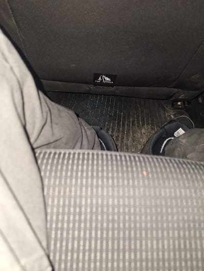 The image shows a car interior. The perspective is from within the car, looking down. A car seat with a grey and white patterned upholstery is visible on the right. To the left is a dark car seat with a "TOP TETHER" tag on it. On the floor, in between the seats, is a black rubber floor mat. A person wearing grey trousers and black shoes with white accents is also visible. No information to indicate if this image was taken in the city of Boliartsi, Bulgaria, exists.