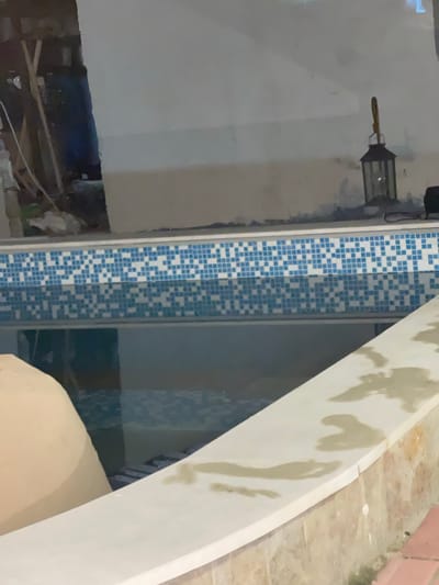 The image displays an indoor or semi-enclosed swimming pool area located in Debelt, Bulgaria, under dim illumination. The pool features a waterline adorned with a mosaic pattern of small blue and white tiles, and its surface reflects the surrounding environment. A white coping borders the pool in the foreground, showing several distinct wet marks from water splashes. In the lower-left quadrant, a large, light-colored, curved object is partially visible within or adjacent to the pool water. The background consists of unfinished, light-grey walls, with what appears to be construction debris and wooden elements visible on the far left. On the right side, a dark-colored metal lantern with a top handle is positioned on a surface above the pool's edge. No individuals are present within the scene.