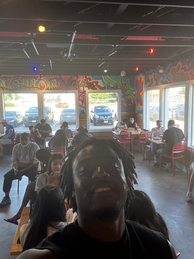 A selfie of a person with locs smiling, likely inside a restaurant. Several other people are seated at tables in the background. There are windows along one wall with views of a street and parked cars. Graffiti-style artwork decorates the wall above the windows. Exposed ceiling beams and hanging lights are visible. The restaurant appears to be located in Arlington, USA.