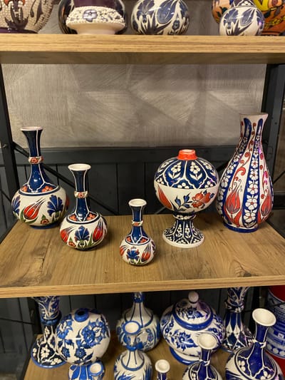A detailed, forensic, matter-of-fact description of the scene:
The image displays a collection of ceramic vases and decorative items arranged on two wooden shelves. The upper shelf holds several larger, ornately decorated pieces, some with purple and white floral patterns and others with blue and white designs featuring leaf-like motifs. Below, the second shelf exhibits a more prominent arrangement of ceramic vases. On the left side of this shelf are three vases of varying heights, all featuring a white base adorned with blue and red tulip and floral patterns. To the right of these vases, a round, stemmed decorative bowl with a red lid is placed, also decorated with blue and white floral designs and red accents. Further to the right is a tall, slender vase with a white base and intricate blue and red floral patterns. The background of the upper shelf features a textured grey wallpaper. The lower shelf is also made of wood and is supported by black metal framing. On this lower shelf, a collection of blue and white ceramic items is visible, including what appear to be vases, a lidded pot, and candle holders, all decorated with floral motifs. The environment suggests an indoor display, likely a shop or a home decor setting. There are no people or actions depicted in the image. The scene is static and focuses entirely on the ceramic display.
The image does not provide any information that allows for determining its location within the city of Kocasinan, Turkey.