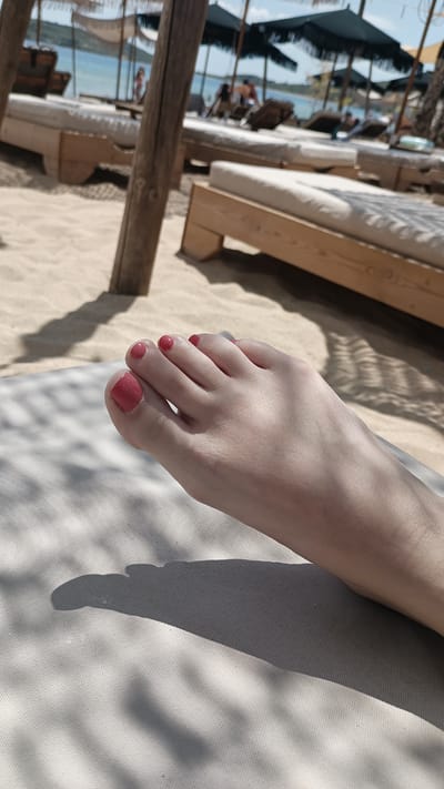 A close-up, low-angle shot focuses on a woman's bare foot, painted with coral-pink nail polish, resting on a white canvas surface. The foot is positioned on the right side of the frame, with toes pointing towards the left. In the background, the sandy beach of Nea Peramos, Greece, is visible, with rows of light-colored wooden sun loungers and beach umbrellas. The sea, a calm blue, is in the distance, with a few people visible on the beach and in the water. Sunlight casts dappled shadows from an overhead structure onto the canvas and the sand.