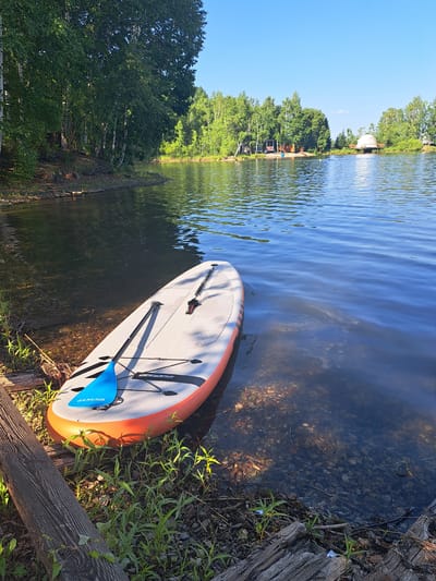 An inflatable stand-up paddleboard, orange and white with a blue paddle, rests partially submerged at the edge of a calm, clear lake. The lake is bordered by a lush green forest with birch trees visible along the bank. Across the water, in the distance, are several rustic buildings and a geodesic dome structure. The sky is a clear, bright blue. The scene appears to be a tranquil natural setting, possibly a recreational area.