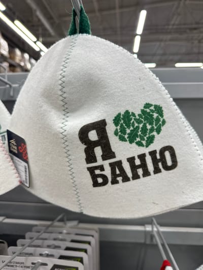 A white, felt sauna hat with green stitching and a green oak leaf heart logo is displayed on a retail shelf. The hat has the Russian text "Я БАНЮ" printed in black, which translates to "I love the bathhouse." The hat is hanging from a metal rod, and in the background, fluorescent lights are visible on the ceiling of a store. The environment appears to be a retail store, possibly a hardware or home goods store, given the packaging of items on the shelves below. The location is identified as Orsk, Russia.