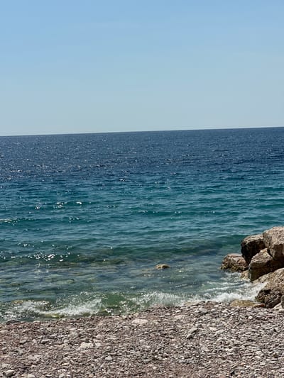 The image depicts a coastal scene at Perazića Do, Montenegro, on a clear, sunny day. The horizon is not visible, as the frame is filled with the water and shoreline. The dominant subjects are the vast expanse of deep blue ocean on the left, its surface shimmering with sunlight. To the right, the water transitions to a clearer, shallower teal hue as it approaches the shore, revealing the rocky seabed beneath. Gentle waves are breaking on a pebble beach composed of numerous small, grey and brown stones. Large, dark rocks are visible along the upper edge of the frame, nestled against the shoreline. A small, indistinct object, possibly a seal or a piece of debris, is visible in the shallow water near the shore. The overall impression is one of a tranquil and natural coastal environment. No text is visible in the image.