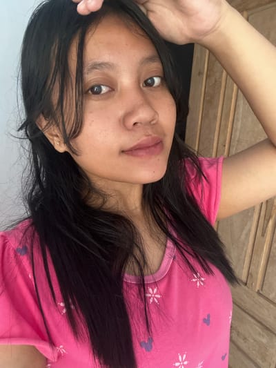 A young woman with long, dark hair is posing for a selfie in Jambearum, Indonesia. She is wearing a bright pink t-shirt adorned with small white flowers and purple Mickey Mouse silhouettes. Her left hand is raised, resting on her head. The background consists of a light-colored wall on the left and a wooden door with vertical panels on the right. The lighting suggests an indoor setting, possibly a home.