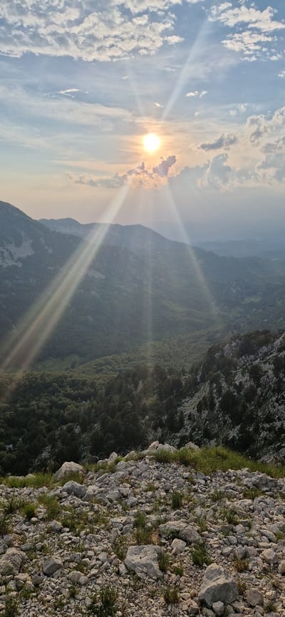 The image captures a breathtaking mountain landscape at sunset, likely in the vicinity of Ubla, Bosnia and Herzegovina. The sun, a bright orb partially obscured by clouds, casts dramatic rays of light across the scene. In the foreground, a rocky terrain is covered with sparse green grass. Beyond this, a lush, verdant valley unfolds, filled with dense trees. Further in the distance, rolling hills and mountains fade into a hazy, atmospheric perspective, bathed in the soft, golden light of the setting sun. The sky is a canvas of soft blues and whites, with scattered clouds that catch the sunlight. The overall impression is one of serene, natural beauty and vastness.