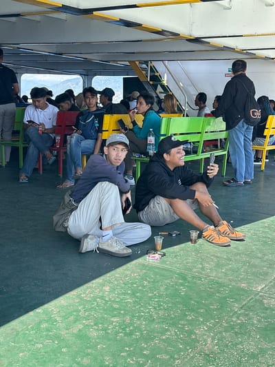Two young men are seated on the floor of a ferry in Ketapang, Indonesia. The man on the left, wearing a dark jacket, light-colored pants, and a baseball cap, is looking to his right. The man on the right, wearing a black hoodie, patterned shorts, and a black baseball cap, is looking at his phone and smiling. Both men have beverages in plastic cups near them. There are other passengers seated on colorful benches in the background, some also looking at their phones. The floor of the ferry is green and appears to be worn. The lighting suggests it is daytime.