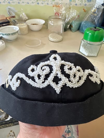 The media file displays a close-up, vertically oriented shot of a black cap adorned with intricate white beadwork. The beadwork forms a swirling, ornate pattern that covers a significant portion of the cap's surface. The cap appears to be made of a textured fabric. In the background, the scene is cluttered with various objects, suggesting a craft or hobby workspace. Visible items include glass jars filled with different types of beads or grains, a green-capped shaker, a small white bowl with contents, and some fabric with a floral pattern. A hand is visible on the right side of the frame, partially obscuring the cap. The lighting suggests an indoor setting, possibly during the daytime. The overall impression is one of a creative project in progress, with the cap being the central focus of attention. No visible text is present in the image. The location context isНовая Тура, Russia.