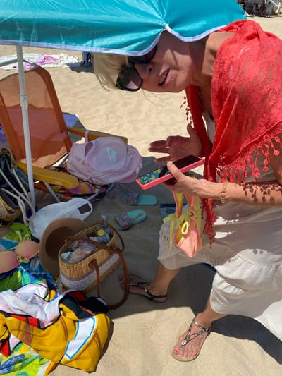 This is a close-up, eye-level, outdoor, medium shot photograph taken on a sandy beach, possibly in Conil De La Frontera, Spain, on a sunny day. The scene is dominated by a woman, likely in her 60s, with blonde hair and sunglasses, looking down at her mobile phone held in her right hand. She is wearing a white dress and a vibrant coral-colored, lacy shawl draped over her shoulders. Her left hand is partially visible, gesturing slightly.
Scattered around her on the sand are various beach items: a turquoise beach umbrella providing shade, an orange beach chair, a pink Nike backpack, a straw hat, a woven basket filled with items, a clear plastic bag containing food items, a bottle of water, and several pairs of flip-flops. Also visible are colorful towels and swimwear, including a pink bikini top and a yellow and pink sarong or cover-up tied with a knot. The sand appears to be fine-grained and light in color, typical of a coastal environment. The lighting suggests a bright, midday sun, casting distinct shadows on the sand.