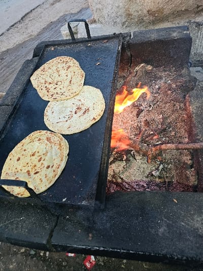 A dark, rectangular grill sits inside a stone oven with flames licking at the wood above. Three flatbreads, speckled with dark brown, are cooking on the grill's surface. The oven walls are made of rough, dark stone, while the fire pit shows ashes and burnt wood. One flatbread is smaller than the other two. A handle is visible on the side of the grill.