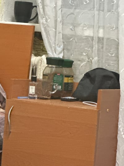 The image depicts a close-up interior scene, possibly a shelf or dresser, within a dwelling. In the background, a white sheer curtain with a decorative pattern is visible, partially obscuring what appears to be a window or another room. On the shelf, several objects are arranged. To the left, a dark-colored mug rests on a white surface. In the center, there are two glass jars, one appearing to contain coffee granules, with green lids. Next to these jars is a clear glass with a handle, and a small glass bottle with a black cap and a label. A dark, soft object, possibly a bag or a piece of clothing, is situated to the right of the jars. A thin blue object, possibly a pen or stylus, lies horizontally on the shelf. In the foreground, a wooden surface with a metallic handle is visible. The scene appears to be domestic and unorganized. There is no indication of the city of Corlăteni, Moldova in the image.
