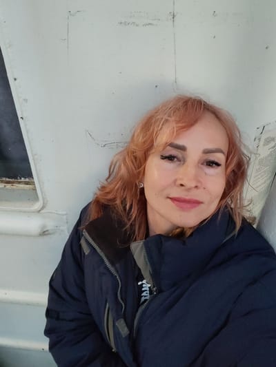 A woman with reddish-pink hair and visible makeup is posing for a selfie in front of a white, textured wall. She is wearing a dark blue jacket with a gray collar and zipper. The wall appears to be an exterior surface, possibly part of a building or structure, with some faint markings and a window frame visible on the left side. The lighting suggests an outdoor environment, possibly during daylight hours. The background is predominantly a plain white wall, with a darker area on the far right that may be a shadow or another part of the structure. There is no information in the image to indicate the specific location within Shcheleiki, Russia.