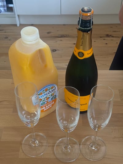 The image displays a bottle of Veuve Clicquot champagne, a jug of Eastcoast orange juice, and three empty champagne flutes arranged on a wooden surface. The items suggest a mimosa preparation. The champagne bottle is green with a yellow label, while the juice jug contains a bright orange liquid. The flutes are clear and have a classic champagne flute shape. The setting appears to be a kitchen counter or table.