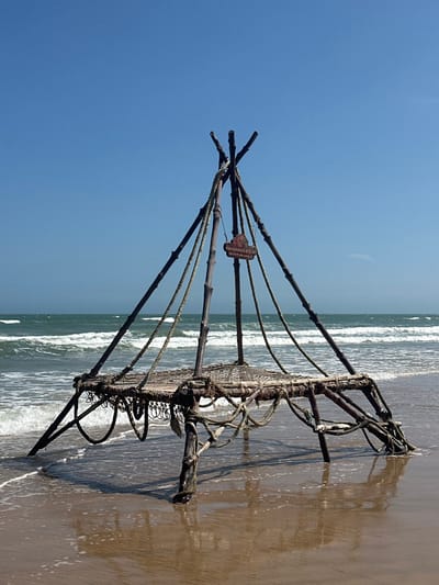 On a sandy beach, a tilted image displays a wooden fishing structure. It consists of several poles converging at a central point and attached to a net-like frame. The structure is partially submerged in the shallow water, with waves gently lapping around it. A small sign with the words "Pandanus Resort" is visible near the center. The sky is a clear blue, and the ocean stretches to the horizon.