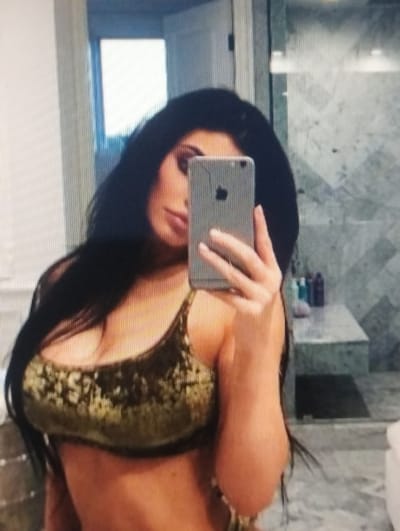 The image shows a woman taking a selfie in a bathroom. She is holding a silver smartphone with her right hand, and her left hand is resting on her leg. The woman has long, dark hair and is wearing a green and gold patterned top. She is looking at the phone and smiling. The background of the image appears to be a tiled wall with a mirror. There is a window with a white blind behind her, and some toiletries are visible on a shelf. The lighting suggests it is daytime. The overall mood of the image is casual and relaxed.