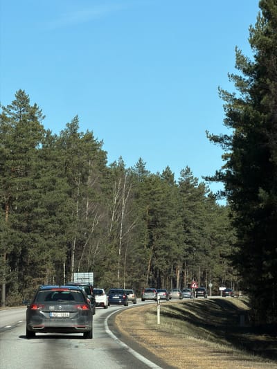 A line of vehicles is stopped on a road in Berģi, Latvia. The road winds through a forested area. The sky is clear and blue. A sign with reflective stripes stands near the roadside. The license plate of the vehicle in the foreground reads "NG-1984".