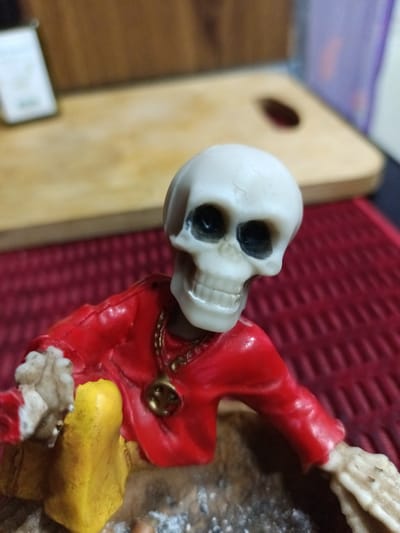 The image shows a close-up of a decorative skeleton figurine wearing a red shirt and yellow pants. The figurine has a gold chain with a circular pendant around its neck. The background is blurred, showing a wooden surface and a red textured mat. There are no discernible details about the location in Yukariyuz, Uzbekistan.