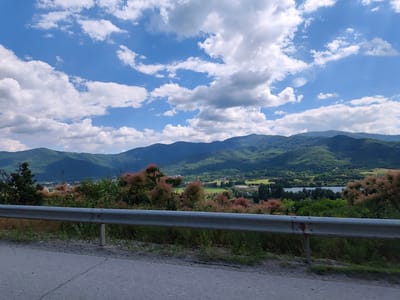 This is a landscape photograph taken from a roadside vantage point, looking out over a valley towards a mountain range under a bright, cloudy sky. In the foreground, a grey metal guardrail runs horizontally across the frame, with a paved road and verge visible below it. Behind the guardrail, a lush green hillside is covered with various trees and bushes, some of which have distinctive fluffy pinkish-red foliage.

The middle ground of the image reveals a valley with a body of water, likely a lake, reflecting the sky. Beyond the water, there are fields and scattered buildings, suggesting a small town or village nestled within the landscape. The distant background is dominated by a range of rolling, densely forested mountains that stretch across the horizon. The mountains are depicted in varying shades of green and blue, with some areas cast in shadow.

The sky is a vibrant blue, dotted with numerous white, puffy cumulus clouds. The lighting suggests it is daytime, likely late morning or early afternoon, given the bright sun and the shadows cast by the clouds and mountains. The overall atmosphere is serene and picturesque, showcasing a beautiful natural landscape. There is no visible text in the image. The location is contextually identified as Pravets, Bulgaria.