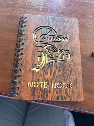 The image shows a wooden notebook with a spiral binding. The cover has a decorative design cut out of it. The word "NOTE BOOK" is cut out vertically on the right side. The notebook is resting on a wooden surface. The location is unknown.