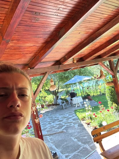 The image depicts a person taking a selfie under a wooden covered patio. The patio has a slatted wooden roof and visible wooden beams. Outside the patio, a lush green garden is visible, featuring a stone pathway, various potted plants, rose bushes with red and white flowers, and a large blue umbrella over a table set with chairs. In the background, a person is seated at the table. The overall setting appears to be a residential outdoor space, likely in Mikre, Bulgaria, given the context. The selfie captures the person's face and upper body in the foreground, with the garden and patio elements occupying the mid-ground and background. The lighting suggests it is daytime, with sunlight filtering through the foliage and illuminating parts of the scene.