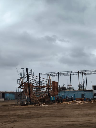 The image depicts a shipbuilding or repair yard in Chimbote, Peru. The scene is dominated by the skeletal framework of a ship under construction, made of wood and metal scaffolding. Several workers are actively engaged in various tasks. Some are gathered around a workbench, possibly cutting or assembling lumber, while others stand nearby.
In the background, a blue industrial building is visible, and behind that, several ships are docked, suggesting a busy port environment. The sky is overcast with heavy, grey clouds, indicating a potentially dull or rainy day. The ground is a dirt or gravel surface, typical of an outdoor industrial setting. The overall impression is one of ongoing work and industrial activity. A yellow triangular warning sign with a black symbol is present near the construction site. The overall mood is functional and industrious, with no overt emotional displays from the workers. The presence of docked ships and the industrial nature of the scene strongly suggest a port or shipyard location.