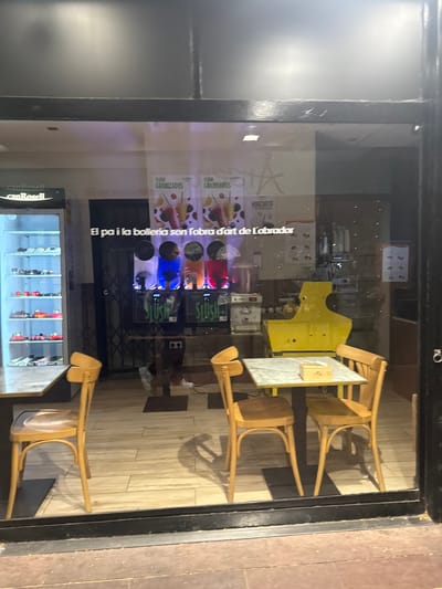 The image shows the interior of a shop visible through a storefront window. The shop appears to be a bakery and beverage establishment.
In the background, there are three slush machines, each filled with a different colored liquid: blue, yellow, and red. Signs above the slush machines read "SLUSH GRANIZADOS" and "HORCHATA". One sign on the wall in the middle of the shot reads "El pa i la bolleria son l'obra d' Obrador". A refrigerated display case to the left is filled with various pastries and cakes. To the right, a yellow machine, possibly a coffee machine or ice cream maker, is visible.
In the foreground, there is a marble-topped table with two wooden chairs. On the table, there is a small wooden box. The floor is tiled with a wood-grain pattern. Reflections on the glass indicate the shop is located on a street, with buildings and a dark sky visible.
Based on the sign "Can Rosell" visible on the refrigerated display case and the text "El pa i la bolleria son l'obra d' Obrador" which is in Catalan, the establishment is likely "Can Rosell" located in Sant Boi de Llobregat, Spain. The name "Can Rosell" is a known business in Sant Boi de Llobregat.