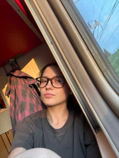 A young woman with brown hair and glasses is looking directly at the camera. She is wearing a gray t-shirt. A red and gray plaid shirt is hanging behind her. The scene appears to be inside a train car, with sunlight streaming in through the window. The background outside the window shows power lines and trees, suggesting a rural or suburban setting. The image's metadata indicates it was taken in Povarovo, Russia.