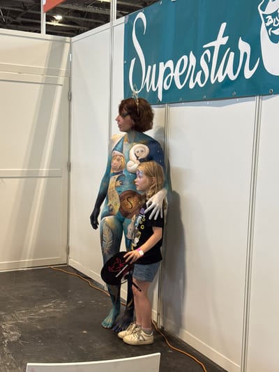 In Villepinte, France, at what appears to be an indoor exhibition or convention center, a person with full-body paint is standing next to a young child. The body paint on the adult features a blue and white design with celestial elements and characters, including moons and what look like stylized figures. A gold, tiara-like accessory is worn on the adult's head. The adult is wearing white gloves. The child, facing to the left, has face paint resembling a butterfly and is wearing a black t-shirt, denim shorts, and sneakers. The child is holding a black fan with a red design. They are positioned in front of a white wall panel and a teal banner with "Superstar" written in white script. The flooring is dark and appears to be a hard surface, with an orange cord running along the base of the wall. The background shows the structure of exhibition booths.