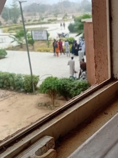This image depicts an outdoor scene in Kwazailewa, Nigeria, viewed from an elevated vantage point through a dirty, light-colored window frame. The immediate foreground shows the window sill, heavily soiled. Below the window, dry, sandy ground with scattered green bushes is visible.
In the midground, a paved walkway or plaza is populated by approximately 10 to 15 individuals, some standing in small groups, others walking. Their attire includes various colors such as orange, maroon, white, and dark blue. A tall light pole, equipped with a solar panel, stands to the left of the walkway, adjacent to a white rectangular sign displaying dark, illegible text. Sections of the paved area are bordered by manicured green bushes.
The background presents a hazy, indistinct landscape featuring trees and distant structures under an overcast or dusty sky. The overall environment suggests a public or institutional outdoor space.