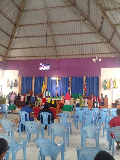 An indoor scene shows a church service in progress, with rows of light blue plastic chairs filled with people. A purple wall forms the backdrop behind the altar area, adorned with a golden cross. Flags or banners hang on the left wall. The building has a high, angled ceiling constructed of metal.