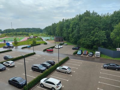 The image shows an outdoor parking lot in Kaluga, Russia, on a cloudy day. The parking lot is paved with interlocking bricks and is lined with low green hedges. Several cars are parked in the lot, including a dark blue sedan, a white SUV, a dark gray SUV, a black hatchback, a silver hatchback, a red hatchback, a black sedan, and a white utility van. In the background, there is a sports complex with a track and field and a basketball court, surrounded by a dense forest. A cell tower is visible in the distance. The overall scene depicts a functional parking area adjacent to recreational facilities and natural landscape.