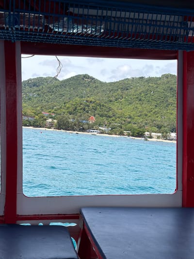 The image depicts a view from inside a boat, looking out through a window. The scene captures a tropical coastline with lush green mountains rising from the sea. A sandy beach lines the shore, with buildings and palm trees dotting the landscape. The turquoise water is calm and reflects the bright sky. The time of day appears to be midday, with clear skies and abundant sunshine, suggesting fair weather. The location is identified as Ko Tao Subdistrict, Thailand, known for its beautiful beaches and marine life.

In the foreground, a blue metal grate is visible, with several bottles of water stacked behind it. The boat's interior is characterized by red and blue painted surfaces, possibly indicating the seating area. The overall impression is one of a serene and picturesque tropical vacation. No specific activities or events are clearly depicted, but the setting suggests a journey by boat along the coast or an arrival at a beach destination. The visible text on the water bottles is partially obscured and difficult to read.