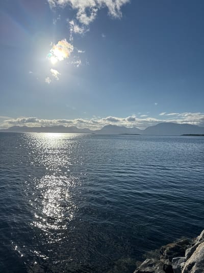 The image captures a serene seascape with a majestic mountain range in the distance, likely a view of the Harstad region in Norway, given the characteristic fjords and mountains. The sun is high in the sky, casting a bright, shimmering reflection on the deep blue water. A few scattered clouds drift across the clear blue sky, one of which is illuminated by the sun, creating a subtle iridescence. The foreground features a rocky shoreline, providing a grounded contrast to the vastness of the sea and sky. No people or immediate actions are depicted, presenting a tranquil and natural landscape.