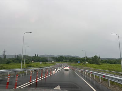 The image captures a view of a wet highway in cloudy weather, likely in the rural area of Lesi Community, Georgia. The road is bordered by metal guardrails and reflective orange traffic cones. A white car is visible driving away from the viewer on the right lane. In the distance, green hills and trees form the backdrop, with power lines and streetlights lining the highway. The overall atmosphere is overcast and damp, suggesting recent or ongoing rain.