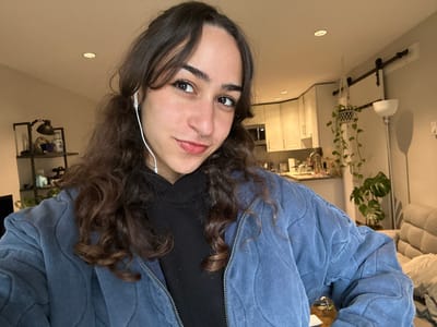 A young woman with curly brown hair poses indoors. She is wearing a blue quilted jacket and a black turtleneck. A silver hoop piercing is visible in her septum. She has earphones in. Behind her is a kitchen with white cabinets and stainless steel appliances. A potted plant is hanging near a pipe fixture. The view is upside down, with the floor appearing as the ceiling.