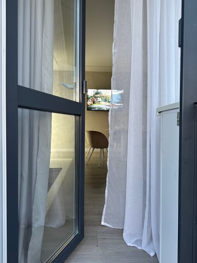 The image shows a view through a large window or glass door from inside a room, looking out. The foreground is dominated by the dark gray frame of the window and sheer white curtains that hang in soft folds. Beyond the curtains, a television screen is visible, displaying a landscape scene with what appears to be a small village or settlement and trees. To the right of the TV, a wooden chair with a metal frame is partially visible, suggesting an indoor setting. The floor visible through the glass is light wood paneling. The overall impression is of a modern interior with a view of an external scene. The lighting suggests it is daytime, and the scene appears to be calm and still. The location is identified as Nizhneyarkeyevo, Russia. There is no discernible text in the image.