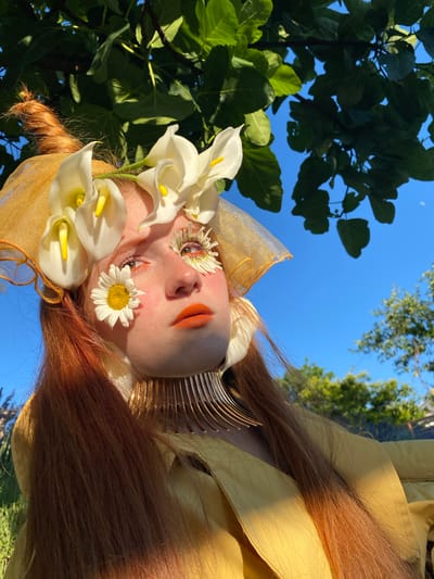 The image shows a close-up, vertically oriented portrait of a person adorned with flowers. The individual has striking makeup, including orange eyeshadow and lipstick, and small white flowers are placed on their eyelids and cheek. White calla lilies are arranged around the person's face, and a daisy is positioned on their cheek. The subject has long, reddish-brown hair and is wearing a yellow garment and a gold, translucent headpiece. A metallic, spiky accessory is visible around the neck area.

The background features a clear blue sky with hints of trees and foliage, suggesting an outdoor setting. The lighting indicates it is daytime, likely with the sun casting a warm glow, illuminating the subject's face and hair. The overall aesthetic is artistic and ethereal, with a focus on nature and elaborate embellishments. The image evokes a sense of fantasy or whimsical portraiture. No text is visible in the image. The provided location context of Verkhneye Uchdere, Russia, suggests the natural environment may be characteristic of that region.