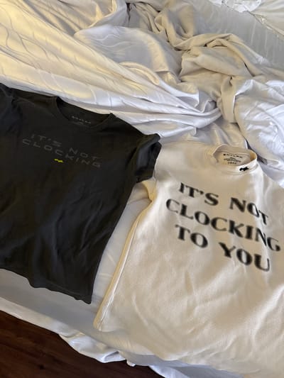 The image shows two t-shirts, one dark grey and one off-white, lying on a rumpled white bed sheet. Both shirts have the phrase "IT'S NOT CLOCKING" printed on them. The brand label "SKYLARK" is visible on the neck of both shirts. The bed appears to be in a residential setting, but there is no definitive evidence to confirm the location within Indian Wells, United States. The photo shows no people, actions, or interactions. The environment is limited to the bed linens and the shirts, with a section of dark wood flooring visible in the background.