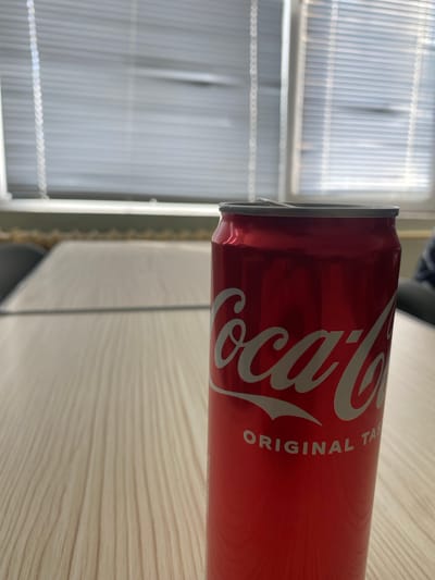 The image features a red Coca-Cola can positioned horizontally on a light-colored surface, likely wood or laminate, which shows a subtle horizontal grain pattern. The iconic white "Coca-Cola" logo is prominently displayed on the can's side, along with the white, uppercase text "ORIGINAL TASTE" below it.
In the background, a window with light-colored venetian blinds is visible, allowing diffused daylight to illuminate the scene. The blinds create a striped effect with vertical lines and horizontal elements. The lighting suggests it is daytime, with soft, natural light coming from the window. The setting appears to be indoors, possibly a room in Chirpan, Bulgaria, given the location context. There are no people or specific activities depicted; the scene is static, highlighting the beverage can within its indoor environment. The can is in sharp focus, while the background is slightly blurred.