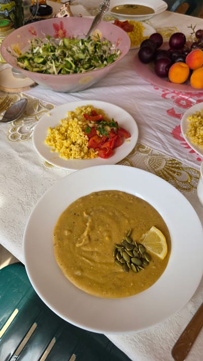 A table is set with a meal, featuring several dishes. In the foreground, a white plate holds a bowl of golden-hued soup, garnished with pumpkin seeds and a lemon wedge. To the left of the soup, another white plate is visible with a portion of yellow grains topped with chopped red tomatoes and herbs. Behind these plates, a large pink bowl contains a salad composed of shredded green vegetables and herbs, with a spoon inside. Further back, a pinkish plate displays a selection of dark plums, apricots, and grapes. A white bowl of soup is also visible in the background, and a jar with a yellow lid is present on the left side of the table. The table is covered with a white tablecloth that has a decorative pattern. The scene appears to be indoors, as suggested by the lighting and the presence of a tablecloth, and is likely set in the city of Kupoyarovo, Russia, based on the provided context.