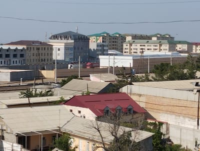 This is an aerial, eye-level, medium-range, wide shot, taken on a clear day, of the city of Margilan, Uzbekistan. The scene depicts a dense urban landscape dominated by residential buildings of varying architectural styles and heights. The buildings feature flat roofs, some with dormer windows, and are constructed from concrete, brick, and stucco. Several buildings are adorned with green, red, and brown metal roofing. A red truck is visible in the middle distance, suggesting activity and transportation within the city. The surrounding environment is a mix of developed urban areas and hints of vegetation, with trees scattered throughout. The sky is clear and blue.