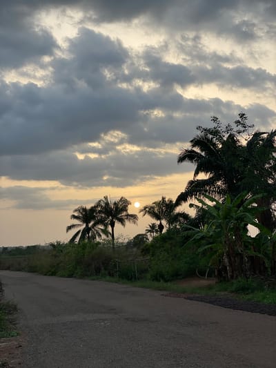 The image depicts a road in Agba Umana, Nigeria, at dusk or dawn. The sky is filled with dark, textured clouds, and the sun is visible, partially obscured by the clouds. Silhouetted palm trees and other vegetation line the side of the road. The road itself is paved and appears to be slightly damp. There are no visible people or other subjects other than the natural elements.