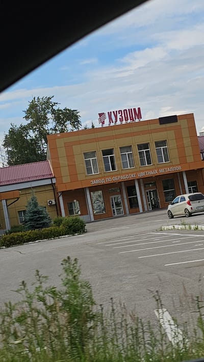 The image shows the facade of a building with a sign that reads "КУЗОЦМ" in red letters. Below this sign, another sign states "ЗАВОД ПО ОБРАБОТКЕ ЦВЕТНЫХ МЕТАЛЛОВ" in white letters. The building has an orange and brown patterned exterior and several windows. To the left of the main building, there is a smaller yellow structure with a maroon roof. In front of the building, there is a parking lot with white lines marking parking spaces. A white car is parked in the lot on the right side of the image. There are green trees and bushes in front of the buildings and tall grass in the foreground. The sky is blue with white clouds. The overall scene appears to be an industrial or commercial property. This is likely the "KUZOCM" plant, which is located in Kamensk-Uralsky, Russia, and is known for processing non-ferrous metals.