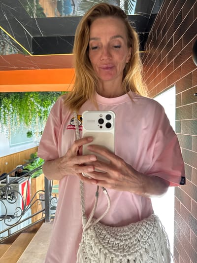 A middle-aged, fair-skinned woman with blonde hair is taking a selfie in what appears to be a reflection in Semenovka, Kyrgyzstan. She is wearing a light pink t-shirt and holding a smartphone with a cream-colored case. A cream-colored macrame bag hangs from her shoulder. The background shows a patterned wall with alternating black and orange sections, and a reflection of greenery and a building with wrought-iron railings. Brown tiles can be seen at the top of the frame.