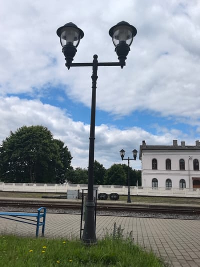The image depicts an outdoor scene, likely at a train station. In the foreground, a paved walkway is visible, bordered by grass with scattered yellow flowers. A blue metal bench is positioned to the left of the walkway. A prominent, dark lamppost with two lights stands in the center, its lights are off. In the middle ground, train tracks run horizontally across the frame. Behind the tracks, a white decorative fence separates the platform from a grassy area and trees. To the right, a light-colored, two-story building with arched windows and a tiled roof is visible, suggesting it's part of the station infrastructure. The sky above is partly cloudy with patches of blue. The overall impression is a tranquil, slightly overcast day at a train station in Лосева, Belarus.
