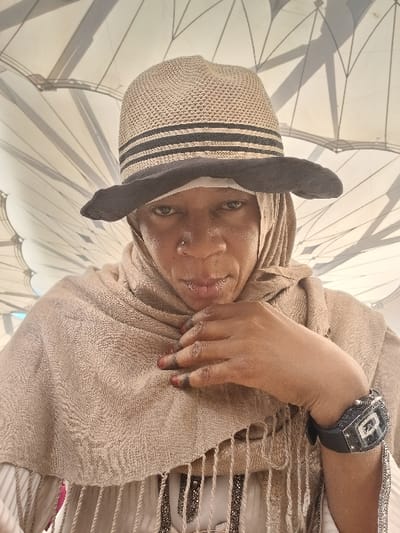 A close-up portrait features an individual, possibly female, with dark skin and a small silver nose stud on the left nostril, looking directly at the camera. The person wears a light brown, woven straw-like hat with two dark horizontal stripes, placed over a light-colored head covering that also drapes over the shoulders and chest. The head covering is textured and has fringes along its lower edge. Underneath, a light-colored garment with visible embellishments is worn. The person's right hand is raised to the chest, with fingers touching the fabric of the head covering. Red nail polish is visible on some fingertips, and dark residue (possibly henna or dye) appears on the nail beds. A large, rectangular black wristwatch with intricate silver-colored detailing is worn on the left wrist. The background consists of the distinctive white, large-scale retractable umbrella structures, identifying the location as the courtyard of the Prophet's Mosque (Al-Masjid an-Nabawi) in Medina, Saudi Arabia.